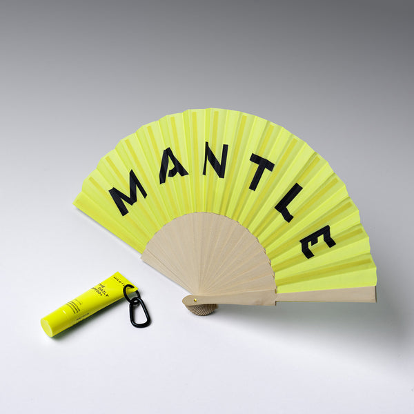 Yellow folding fan with MANTLE text and a yellow SPF tube on a gray background